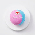Love Shot bath bomb gift set detail showing one pink and blue round bath bomb on a white circular display