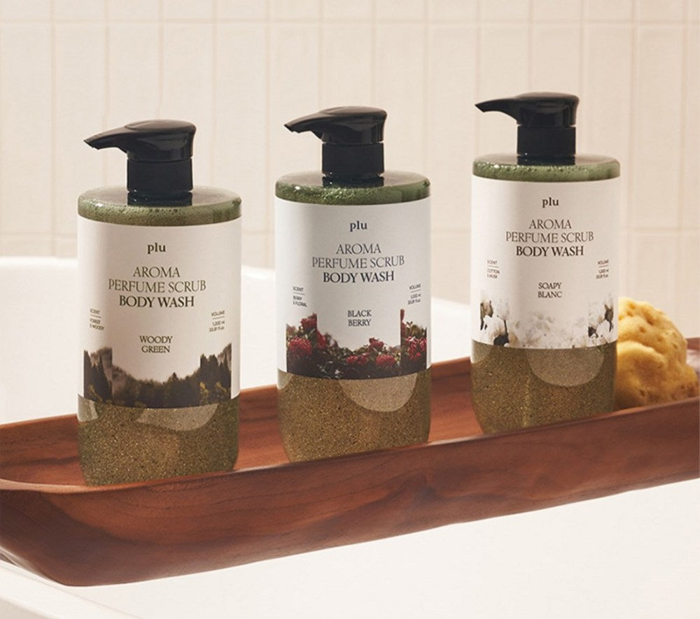 PLU body wash bottles in three scents standing on a wooden bath tray beside a yellow sponge