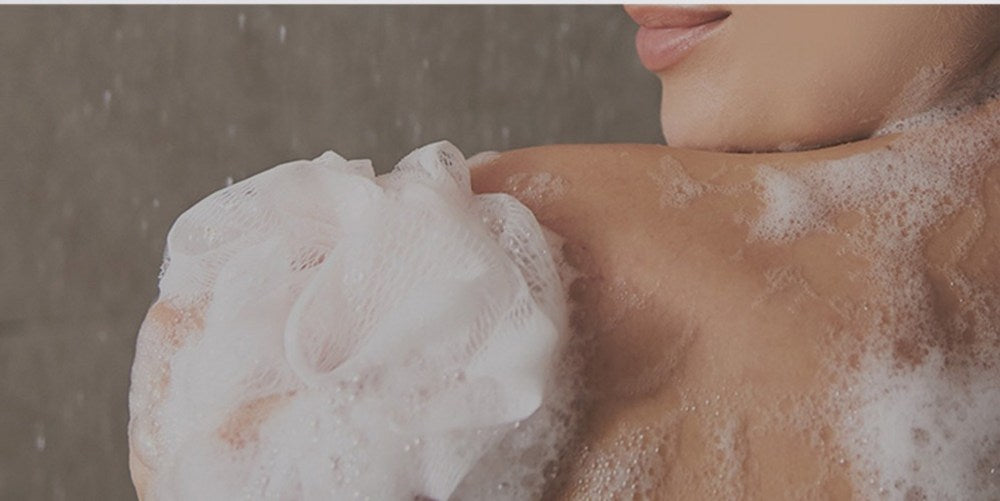 PLU scrub body wash lather on a shoulder, with a white mesh bath puff held against the skin