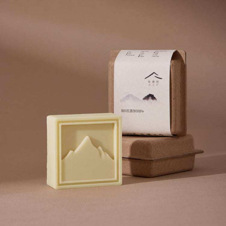 Dooryuheon White Clay Soap pale cream mountain bar beside stacked kraft packaging on a beige background
