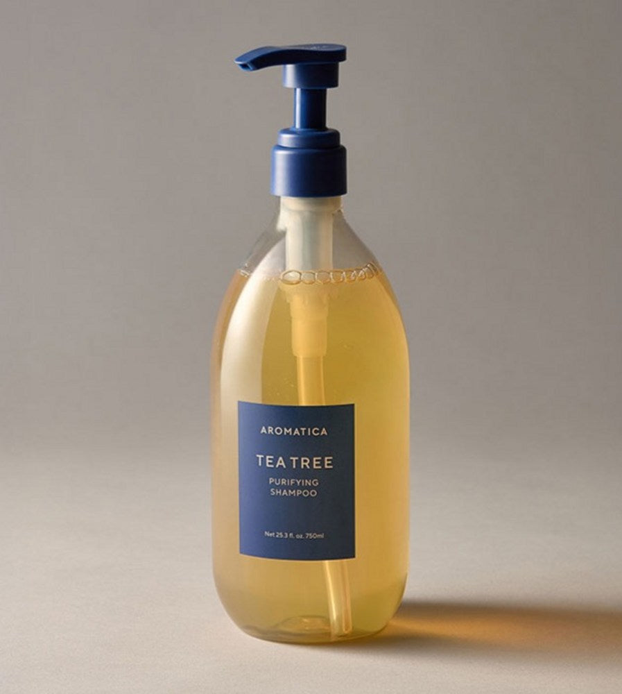 Aromatica Tea Tree Purifying Shampoo with golden liquid and a dark blue pump, standing on a grey studio surface