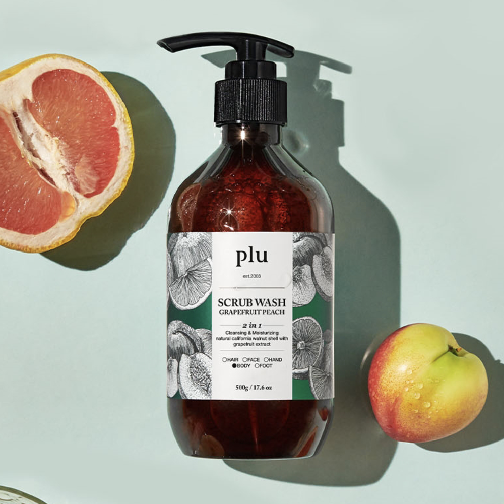 PLU body scrub wash in an amber pump bottle beside cut grapefruit and a peach on a pale green surface