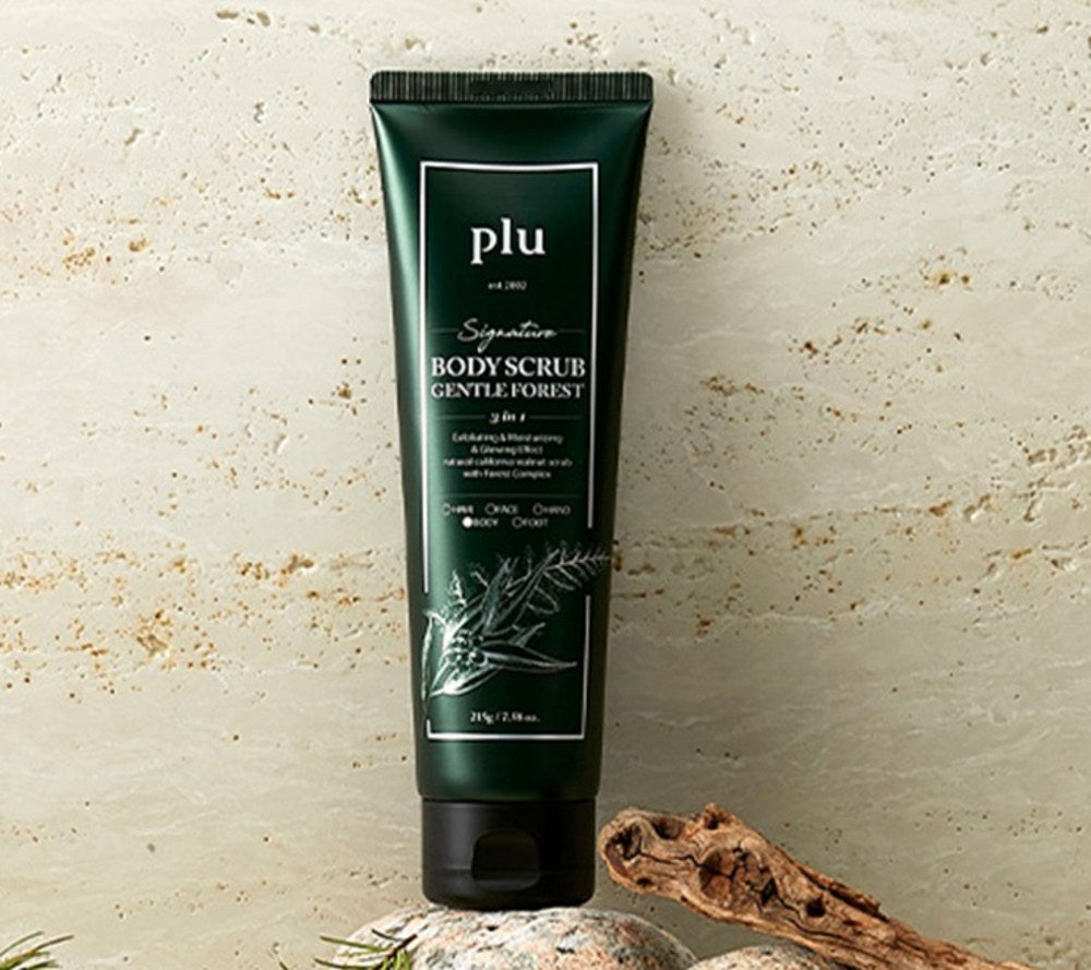 PLU Gentle Forest body scrub tube standing against a pale stone wall with wood and stones below