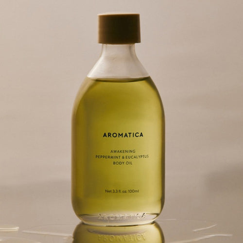 Aromatica Awakening body oil with green-gold liquid and a dark cap, standing on a reflective surface in warm light