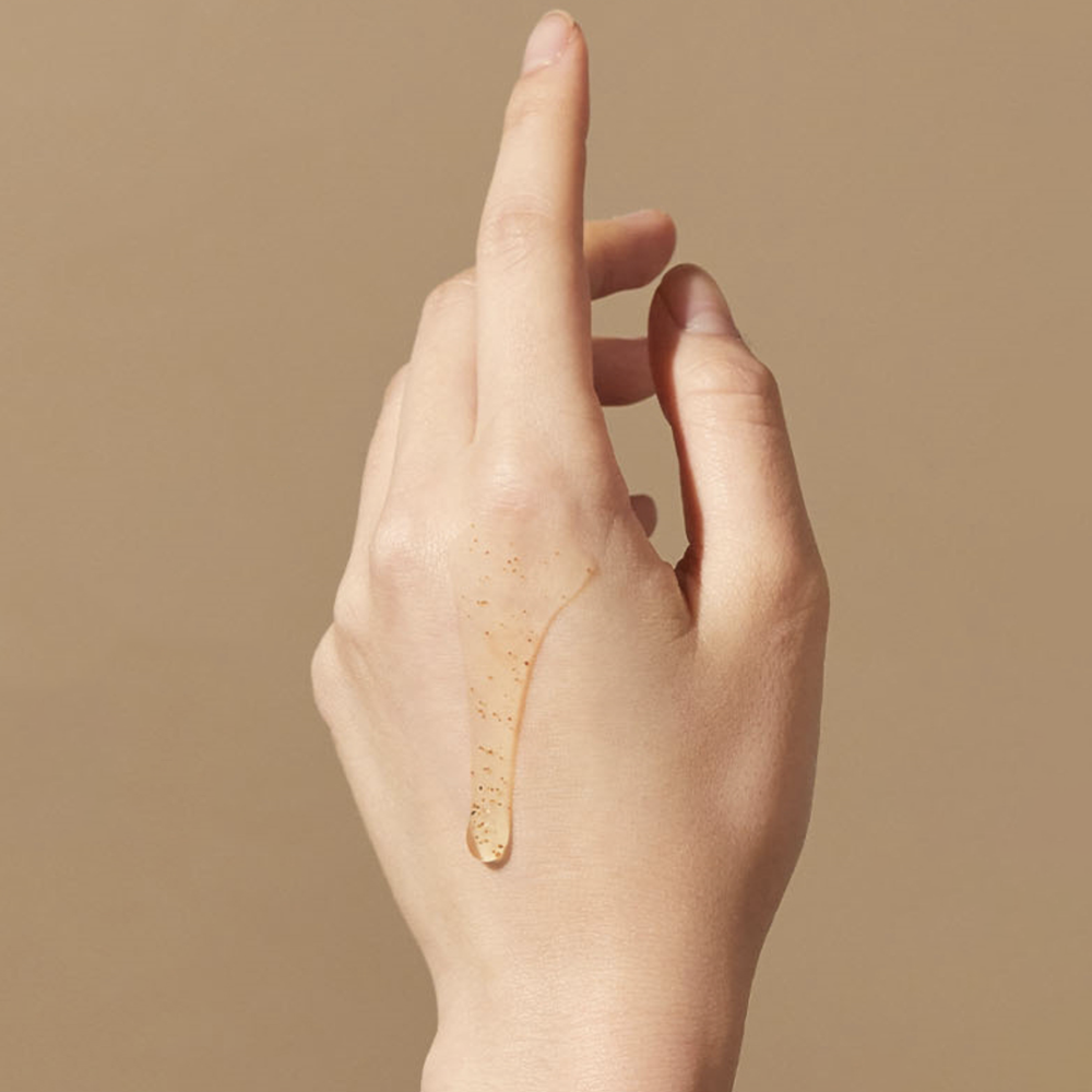 PLU body scrub wash spread across the back of a hand, showing clear gel and scattered scrub particles