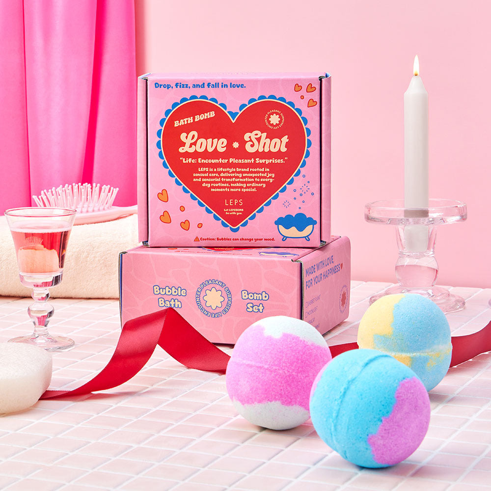 Labsbalm Love Shot bath bombs arranged beside two pink boxes, with a candle, red ribbon and glass on pink tiles