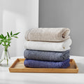 Songwol combed cotton bath towels stacked in beige, ivory, grey and purple on a wooden tray beside greenery