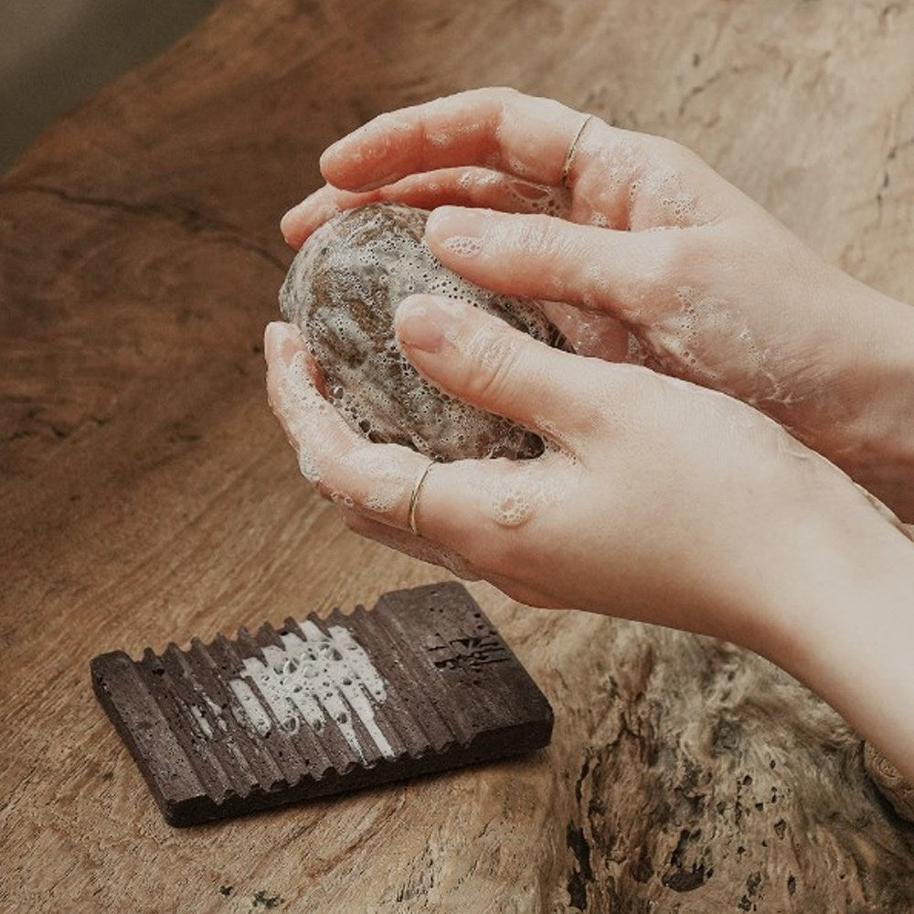 Jeongango extract soap lathered between hands above a wooden surface beside a dark ridged soap dish
