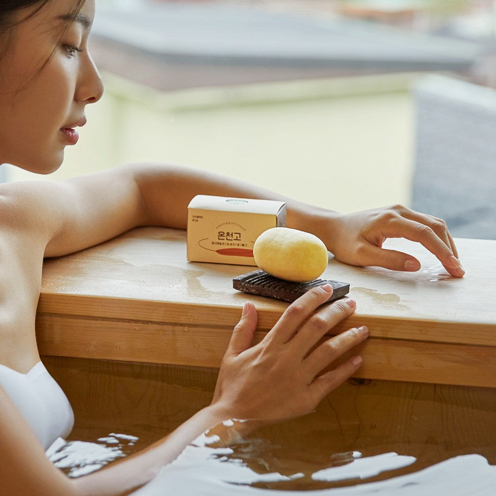 Cheonbinusop sulfur body bar on a dark soap dish beside its box and a bather at a wooden tub