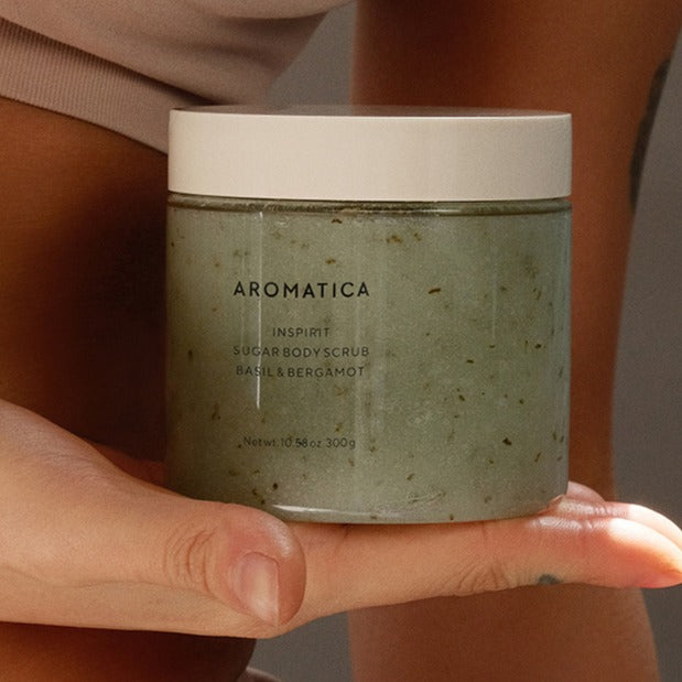 AROMATICA Inspirit Sugar Body Scrub in a pale green 300g jar with a white lid, resting on an open palm.