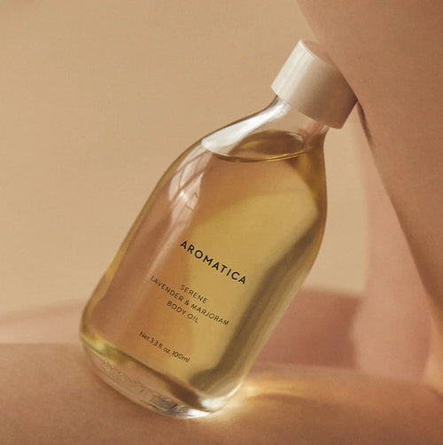 Aromatica Serene body oil tilted against bare skin in warm light, with its 100ml marking and cream cap visible